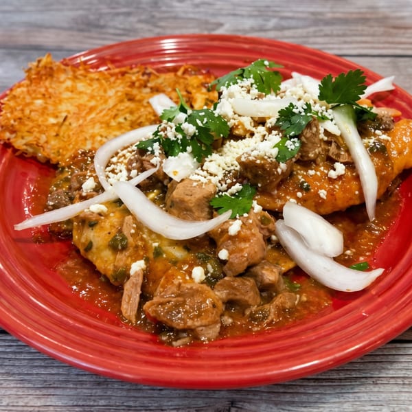 Savory pork stew with onions, cilantro, and cheese, served with a crispy hash brown on a red plate.