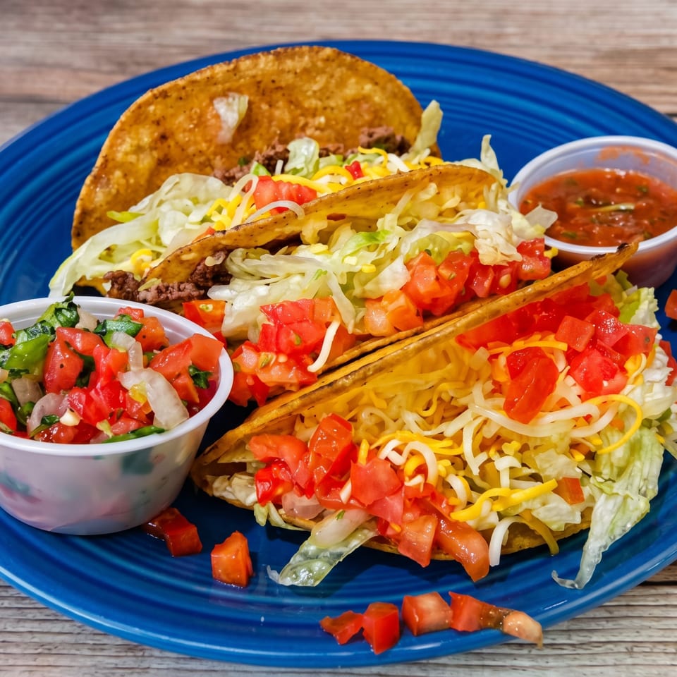 Three hard-shell tacos filled with meat, lettuce, cheese, and tomatoes, served with salsa and pico de gallo.