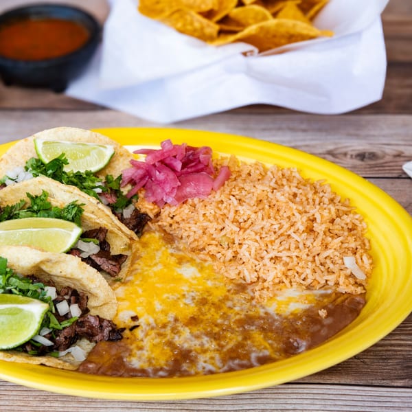 Three street tacos with lime, cilantro, rice, beans, and pickled onions on a yellow plate.