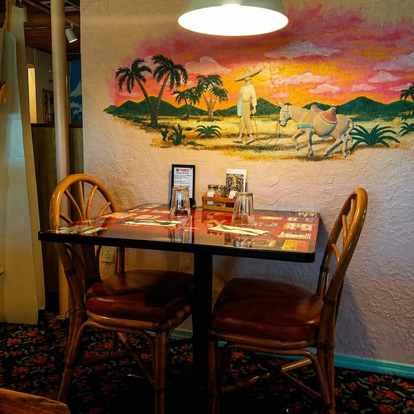 A restaurant table set for two, with a colorful mural of a desert sunset and palm trees in the background.